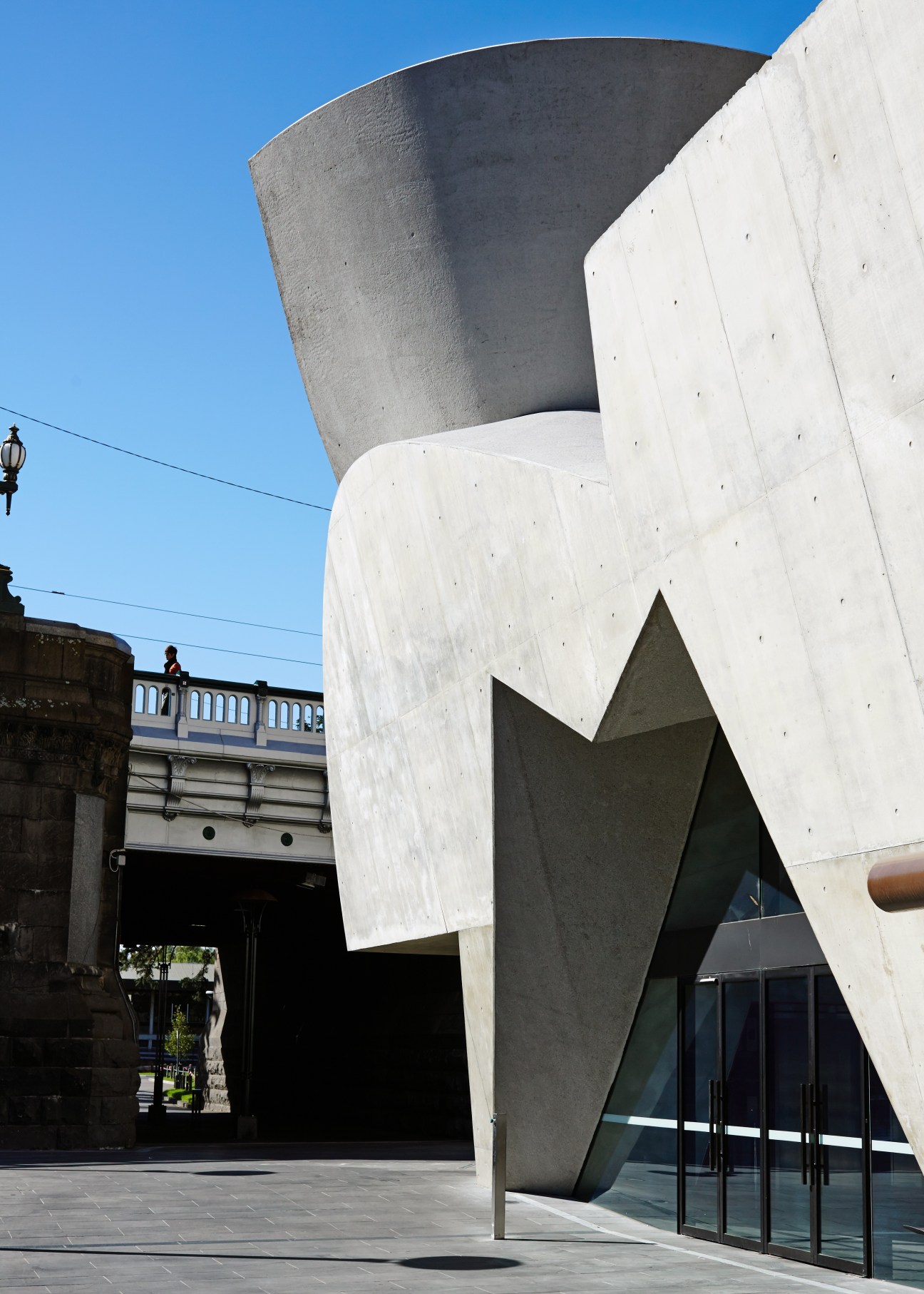Modern white concrete building with curved architectural forms and angular openings beside a heritage stone bridge.