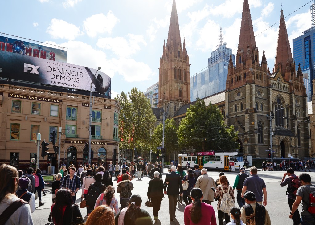 Busy Melbourne street scene with Gothic church spires, heritage buildings, tram, and pedestrians in urban plaza.