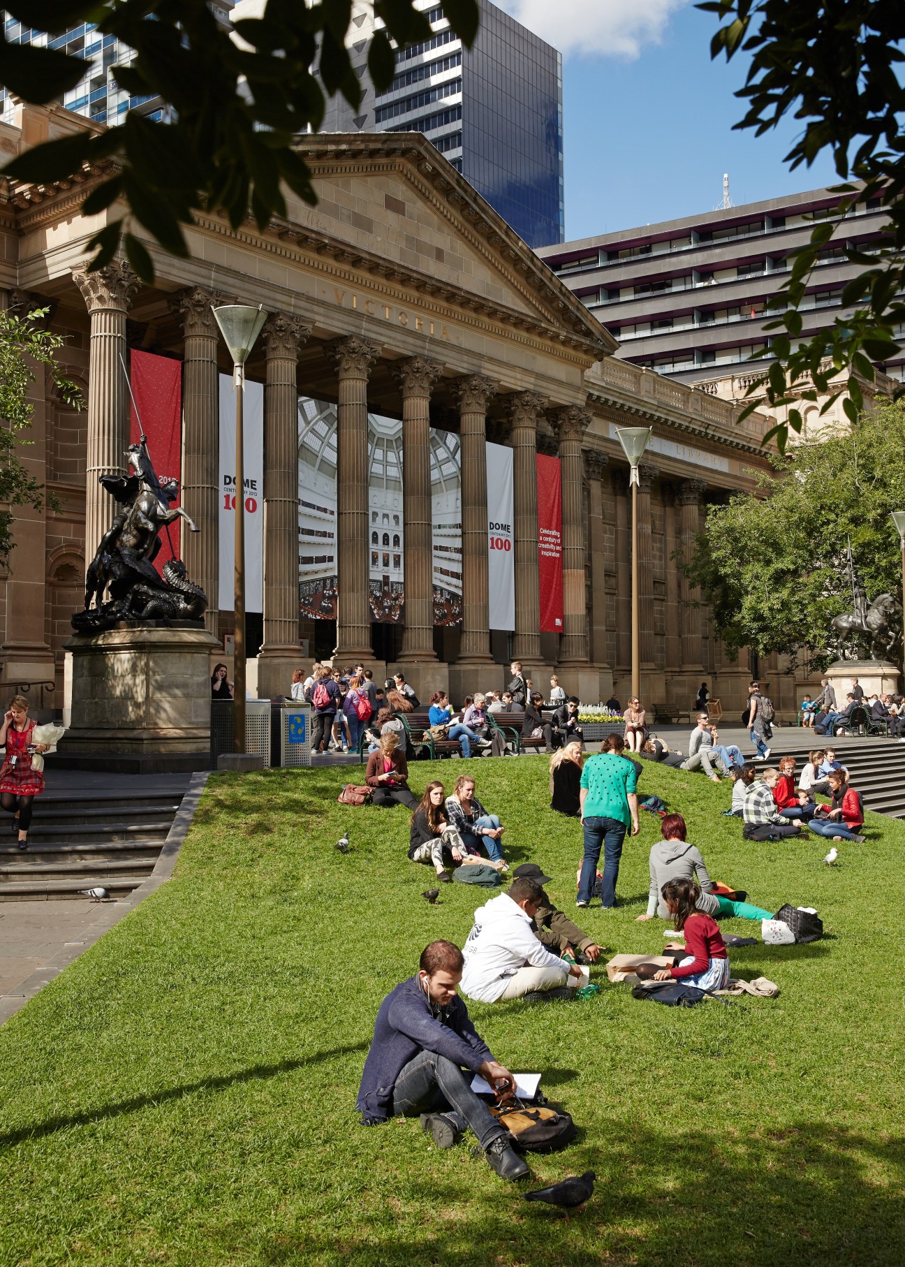 People relaxing on grass in front of a neoclassical building with columns in Melbourne.