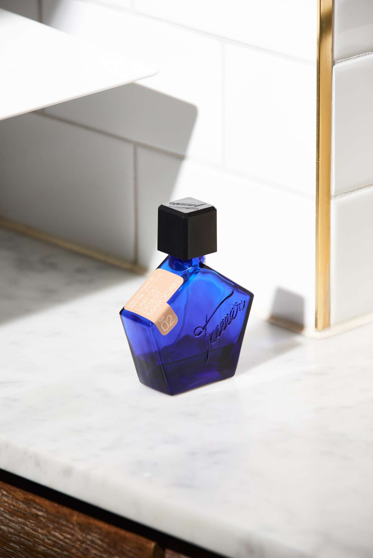 Blue glass fragrance bottle with black cap and tan label on white marble surface.