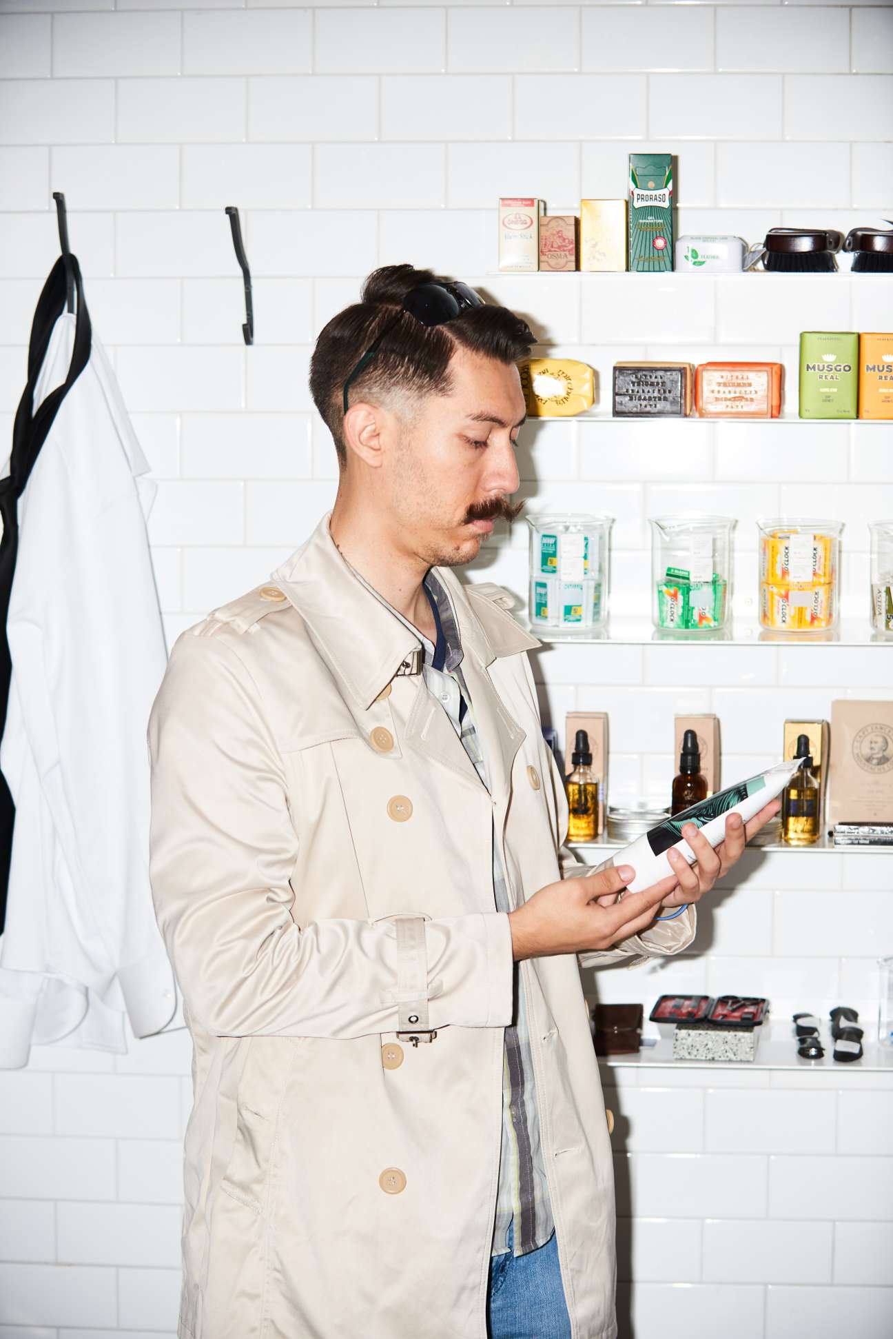 Man with styled mustache examining grooming product at Men's Biz barbershop with white tile wall and shelved products behind him.