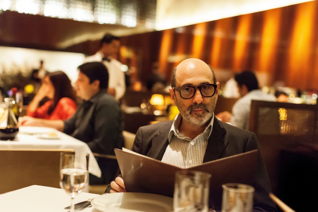 Architect Isay Weinfeld holding a menu at a restaurant table with warm lighting and diners in the background.