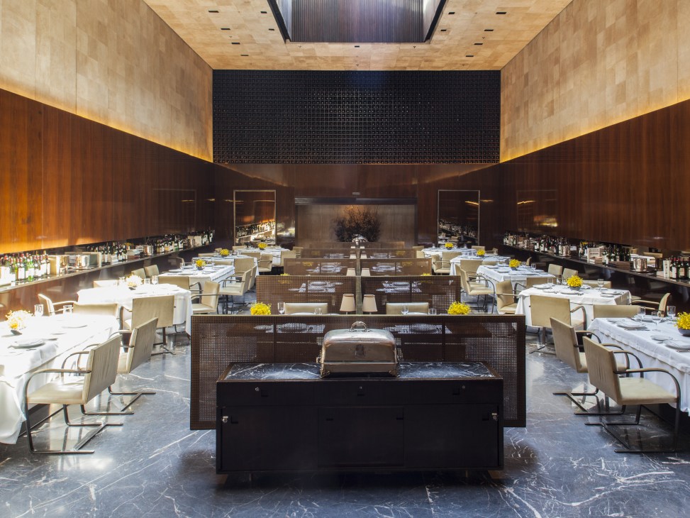 Modern restaurant dining hall with symmetrical layout, wood-paneled walls, cream chairs, yellow flower arrangements, and central service counter.