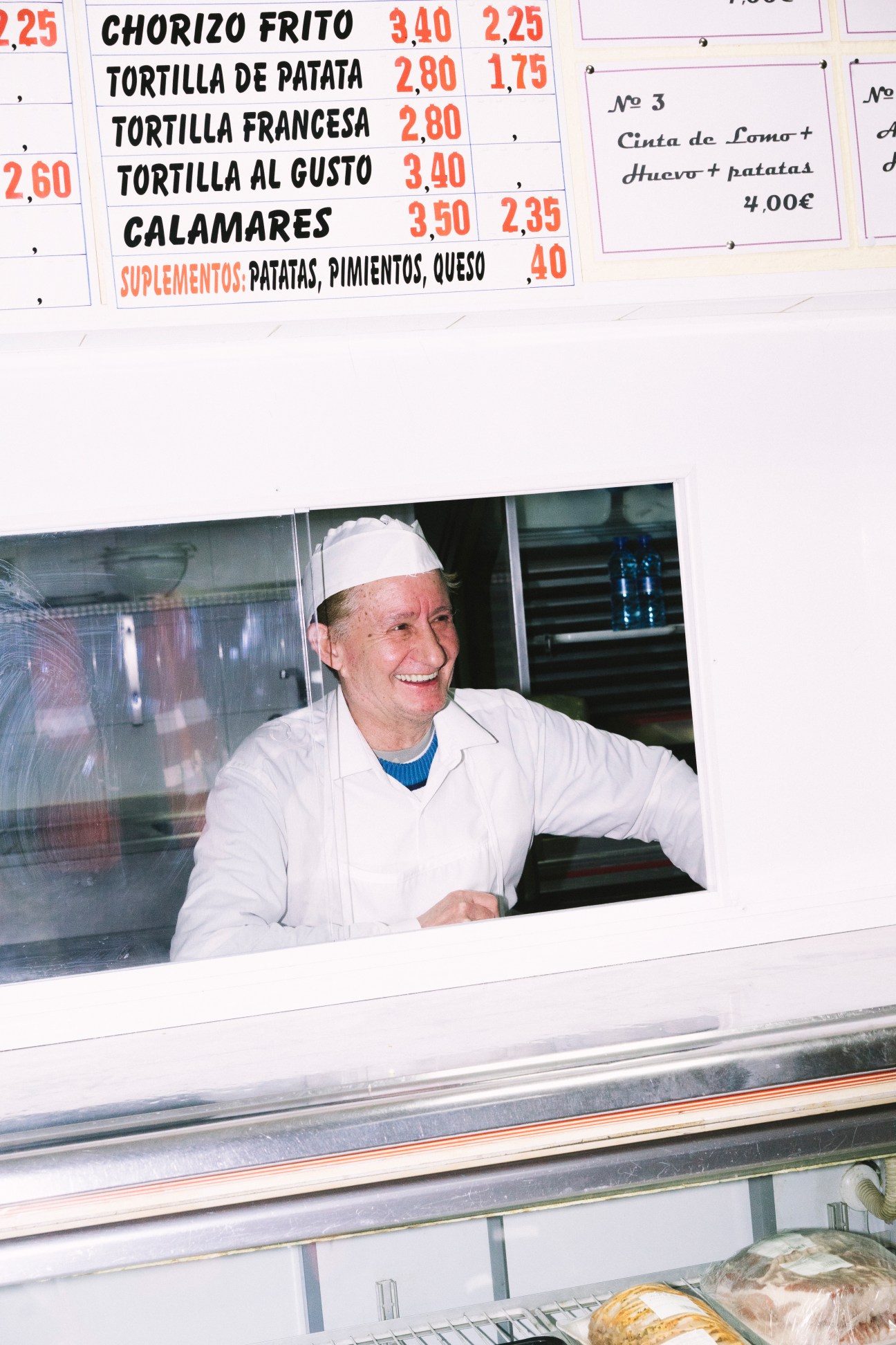 A smiling fishmonger in white chef's coat and headwrap serves customers from a market stall window displaying Spanish tapas menu prices.