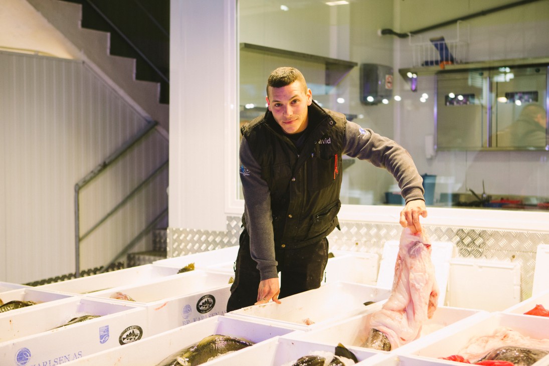 A man in a black jacket holds fresh fish at a stall with white crates at Mercamadrid fish market in Madrid.