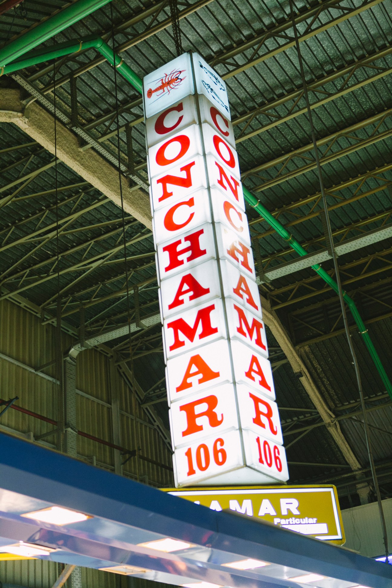 Illuminated vertical sign reading 'LOCHA MAR' with a lobster illustration, hanging in a market building.