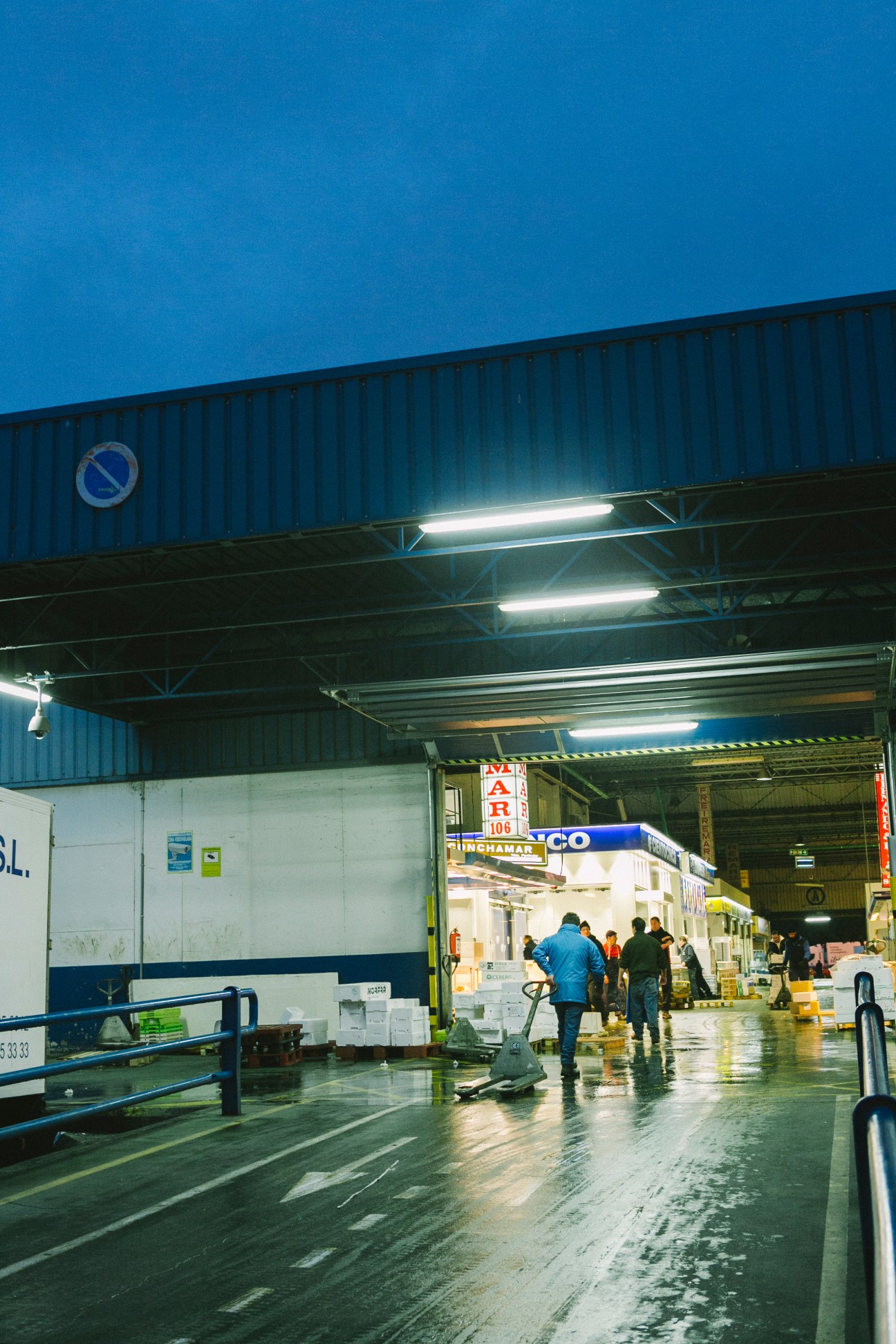 Interior of Mercamadrid fish market at night with wet floor, fluorescent lights, and vendors working under corrugated metal roof.