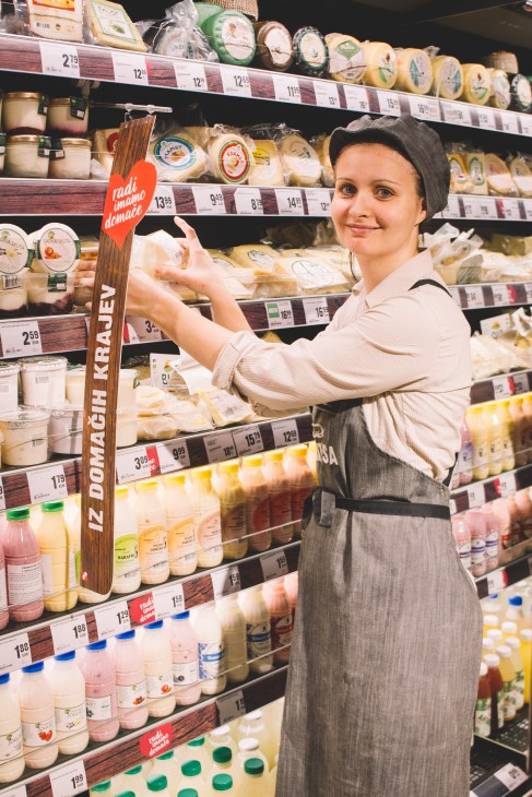 Woman in apron pointing to local dairy products on supermarket shelf marked with red heart logo
