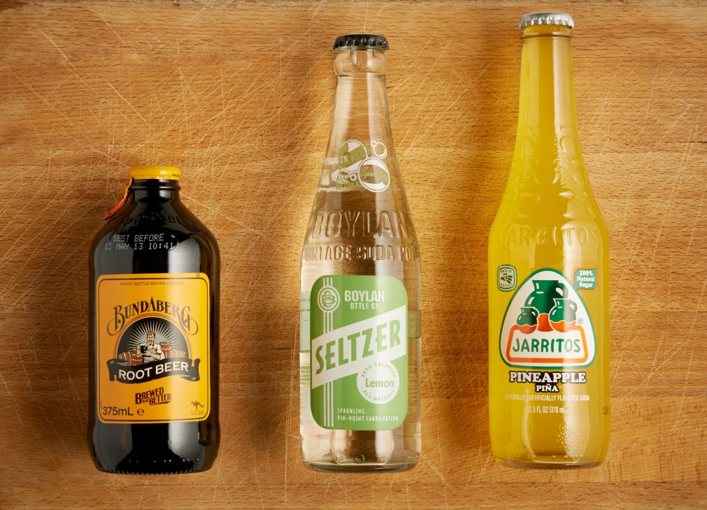 Three soda bottles: Bundaberg Root Beer, Boylan Lemon Seltzer, and Jarritos Pineapple soda on a wooden surface.