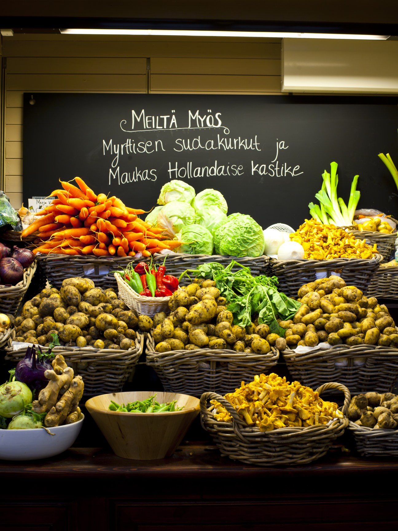 Fresh vegetables displayed in wicker baskets at Merja Valo market stall with Finnish text on blackboard.