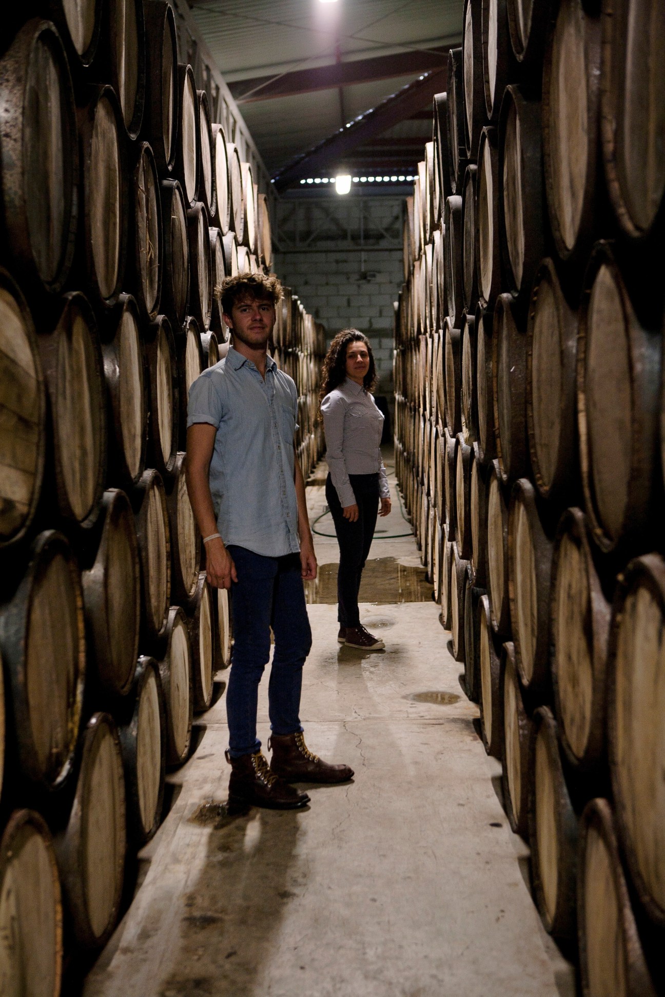 Jorge Rodríguez-Cano and Carmen Casanovas from Mezcal Amores walk through aging barrels in a mezcal distillery.