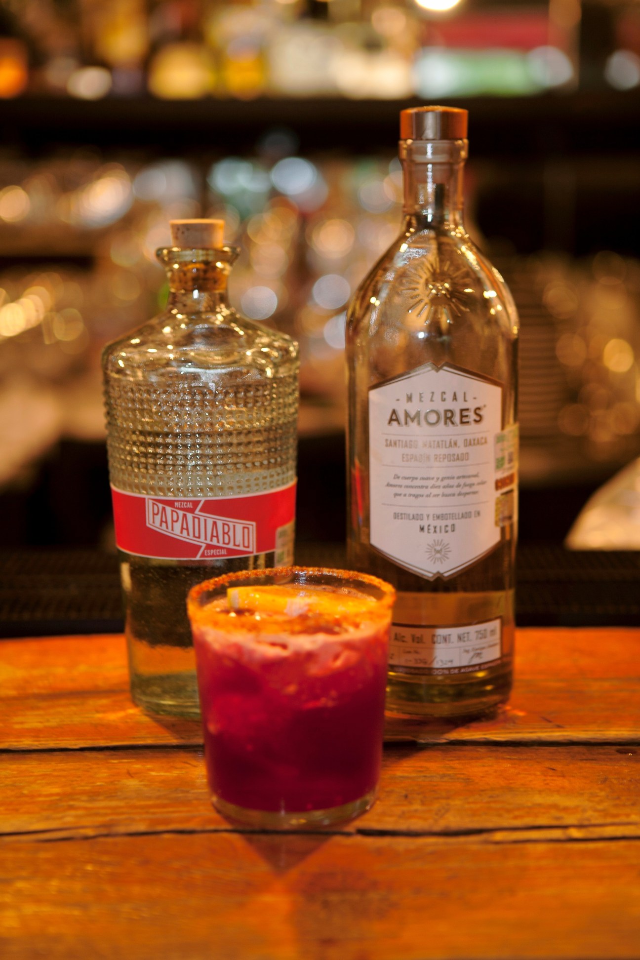 Mezcal bottles including Papadiablo and Mezcal Amores with a cocktail on a wooden bar.