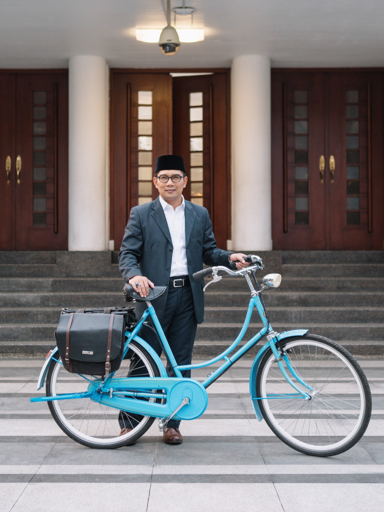 Ridwan Kamil, mayor of Bandung, stands with a blue bicycle in front of a building with white columns and dark wooden doors.