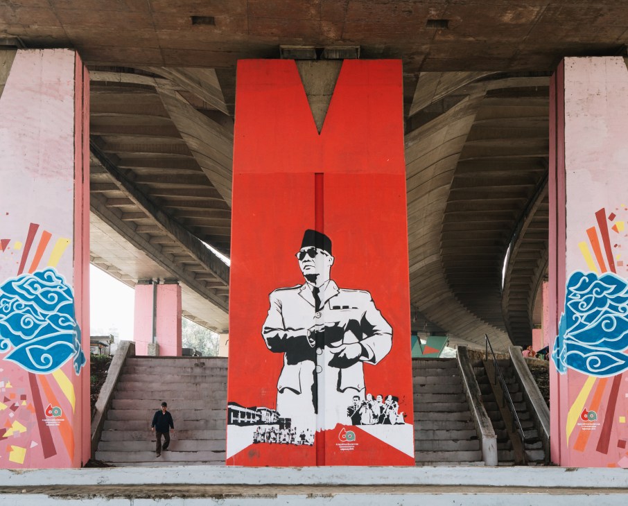 Large red public mural beneath a concrete flyover featuring a figure in military uniform against colorful pink and blue decorative panels.