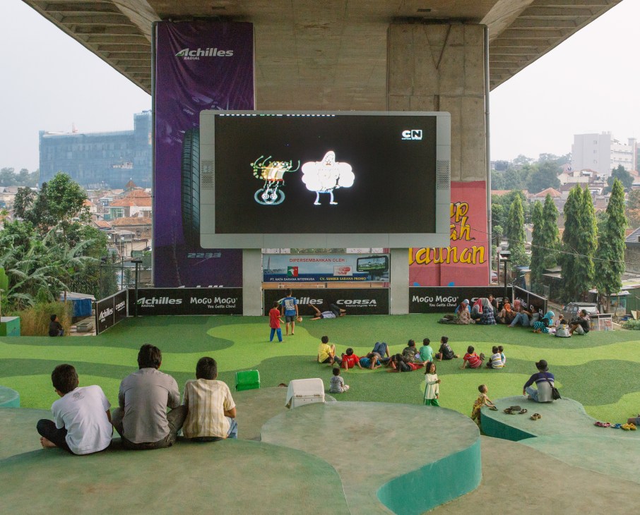 Families and children gather on bright green Astroturf beneath a concrete flyover to watch an animated film on a large LED screen.