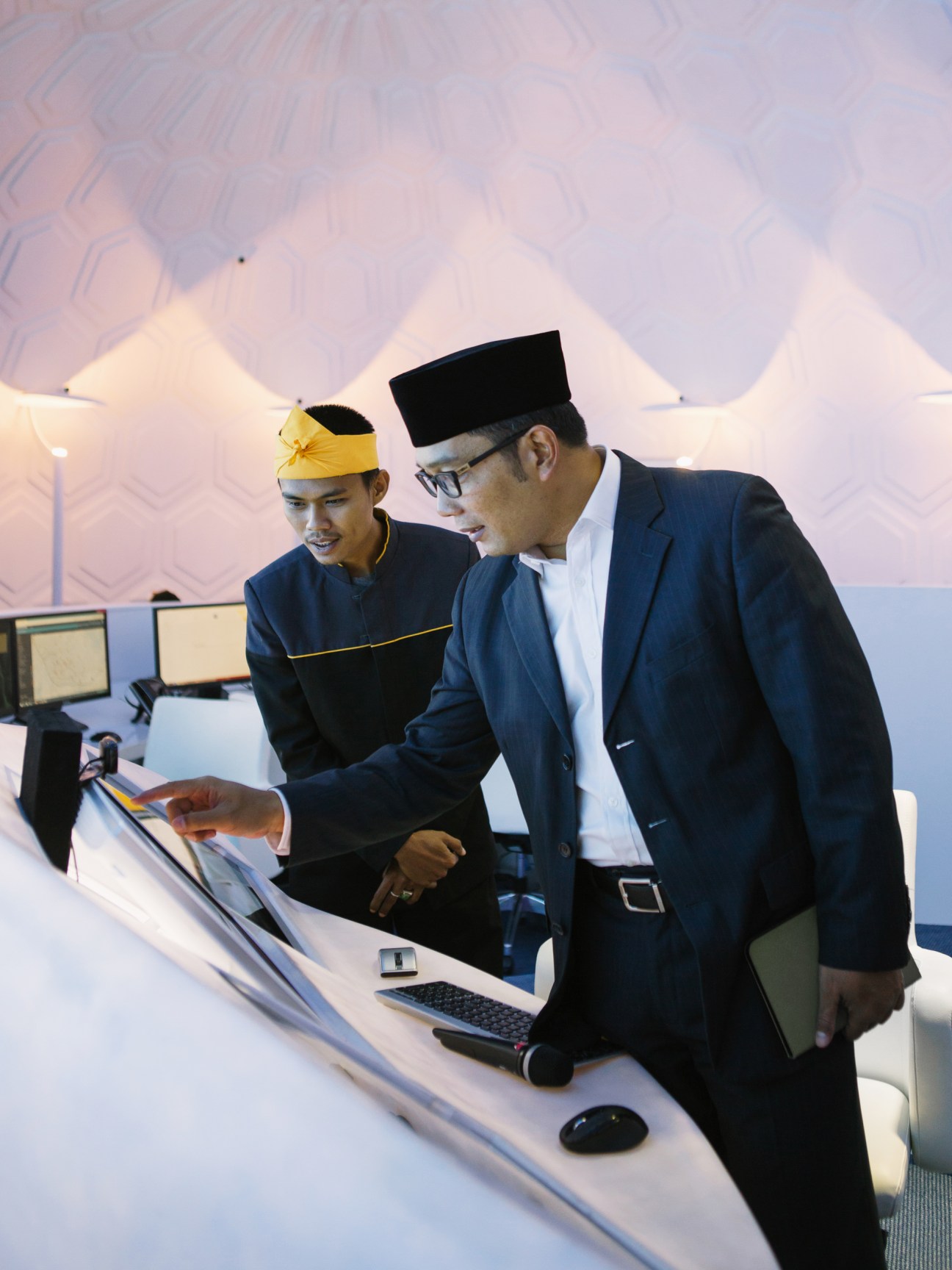 Two officials examine documents at a modern control room console with illuminated geometric ceiling panels.