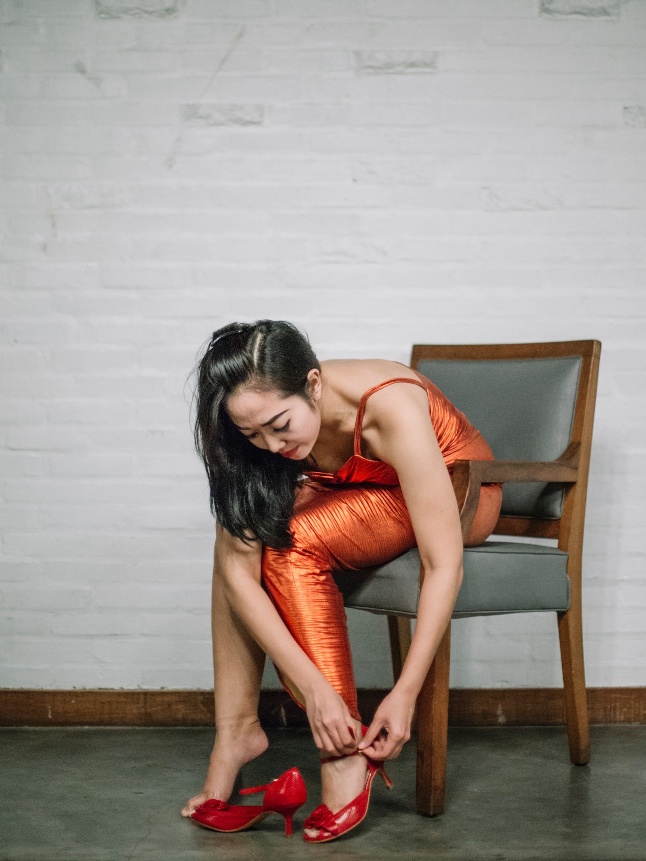 Woman in red dress fastening red tango heels while seated on a wooden chair.