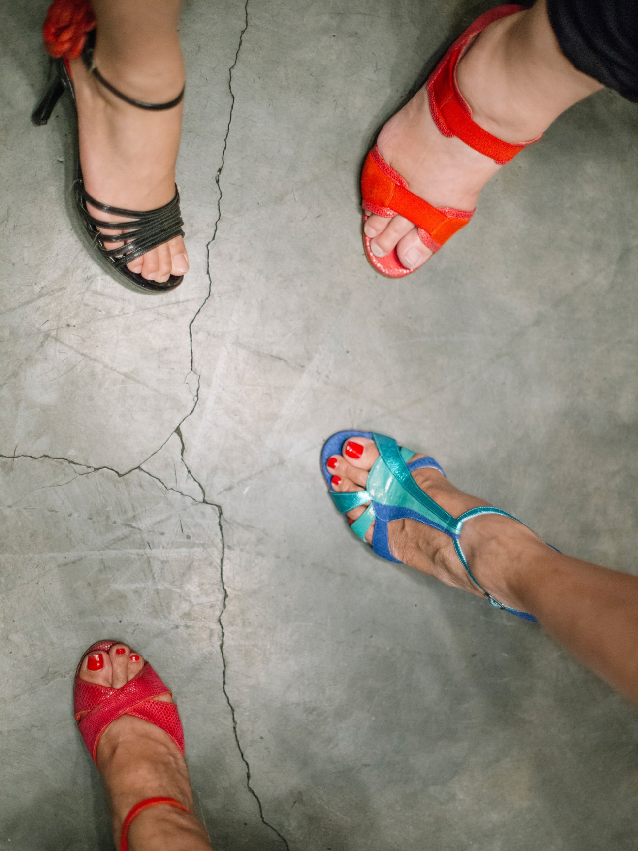 Overhead view of four people wearing colorful Gretaflora tango shoes in red, black, and turquoise on a concrete floor.