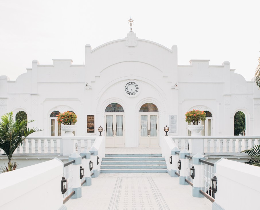 White colonial-style building with clock tower, symmetrical arches, manicured gardens and blue-trimmed stairs.