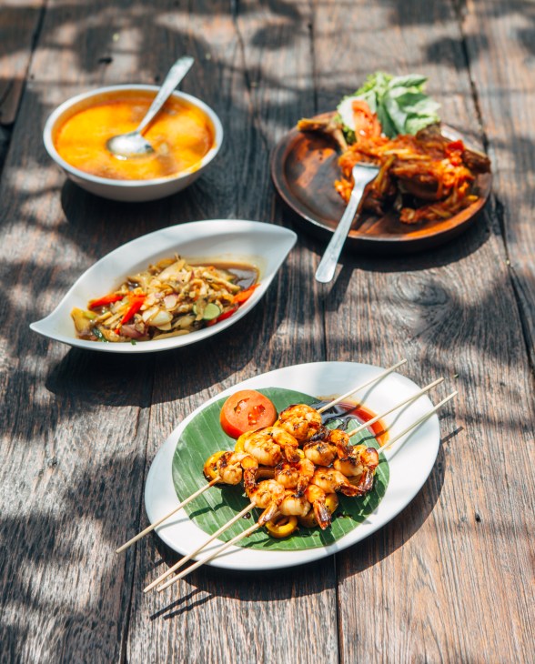 Indonesian dishes including skewered shrimp, soup, and stir-fried vegetables on a wooden table at Dewa-ndaru Culture.