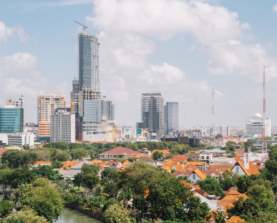 Surabaya's skyline showing modern high-rise construction cranes contrasting with traditional Dutch colonial buildings and orange-roofed houses in the foreground.