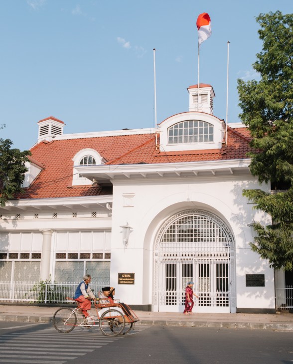 White colonial-style building with red-tiled roof and large arched entrance gate, Indonesian flag flying above, with cyclist and pedestrian in foreground.
