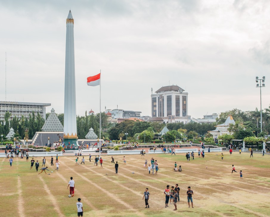 Children and families play in a large open square at Tugu Pahlawan monument in Surabaya, with a tall white obelisk and Indonesian flag visible.