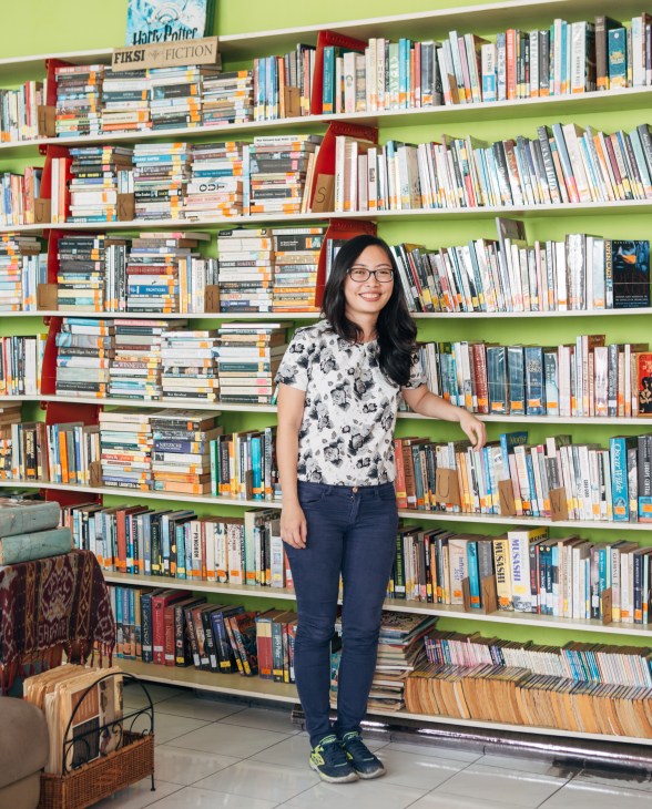 Woman wearing glasses and patterned shirt standing in front of green shelving filled with books in a library.