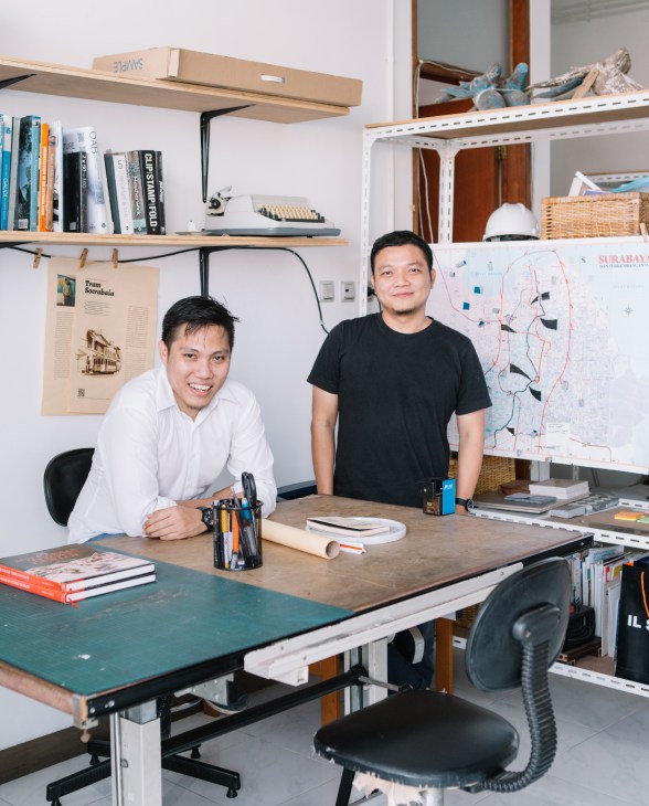 Two men stand at a drafting table in a bright studio space with shelves, books, and architectural plans visible behind them.