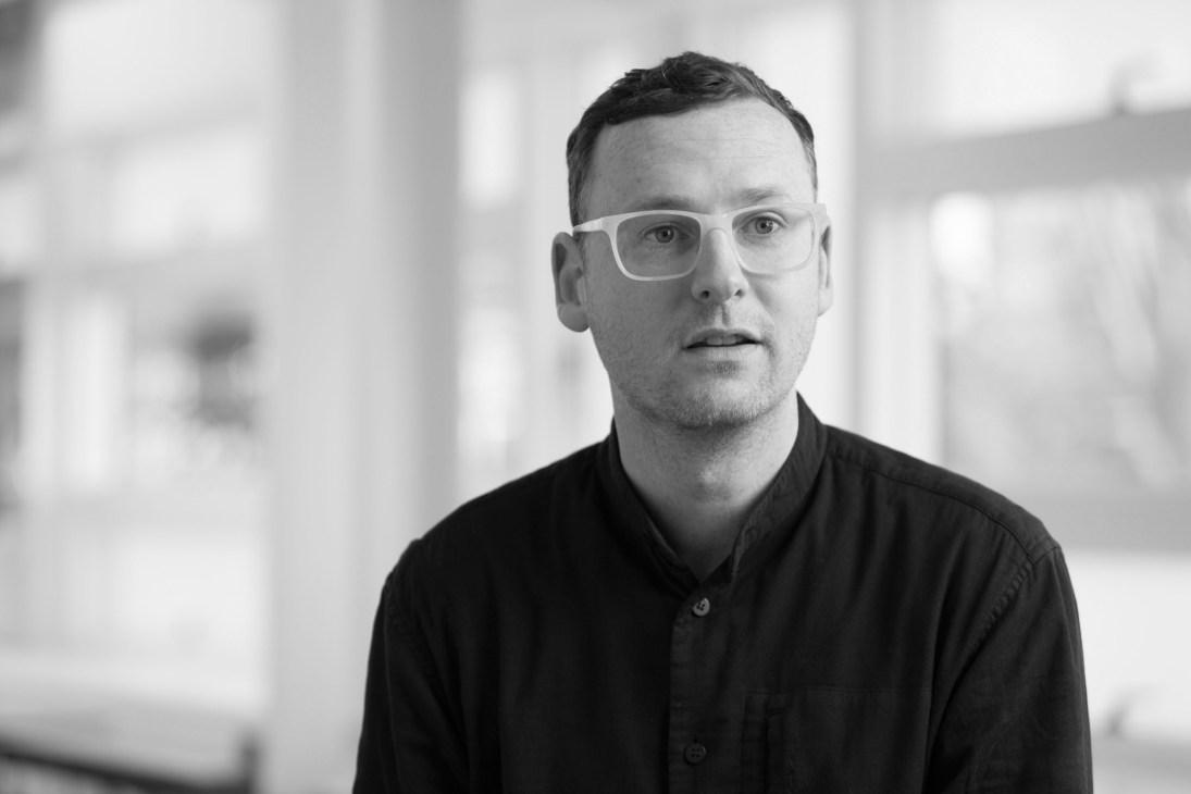Man wearing white-framed glasses and dark shirt in black and white portrait photo.