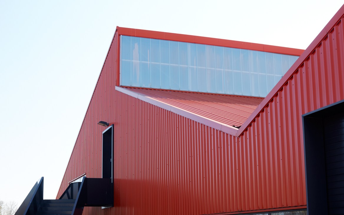 Red-brick factory building with large angular glass window and modern architectural design.