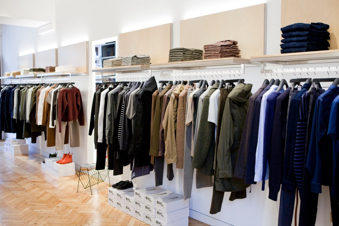 Interior of minimalist MHL store with parquet flooring, hanging garments in neutral and blue tones, and folded items on shelves.