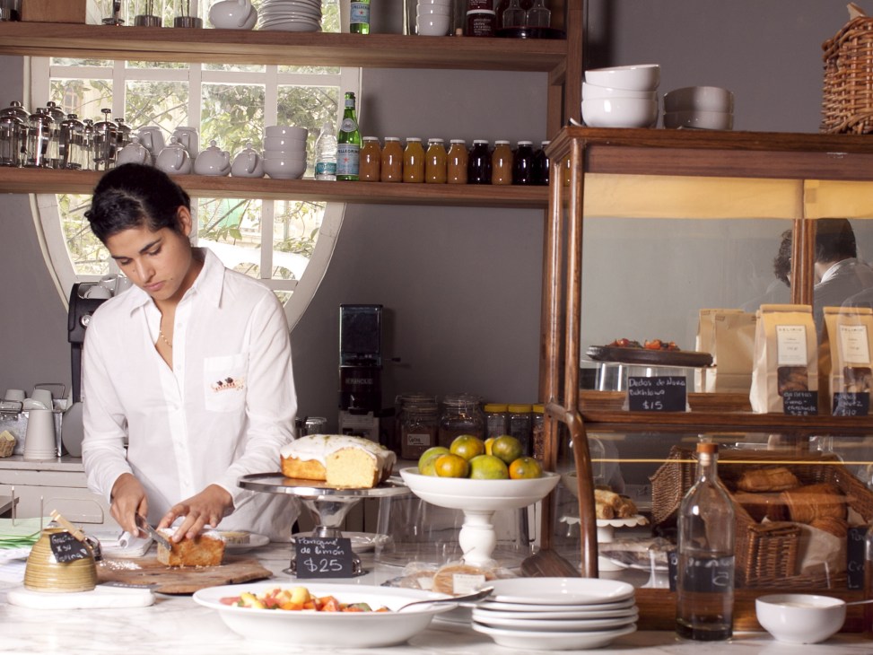 Woman in white shirt preparing food at counter with shelves of bottles and bowls behind her.