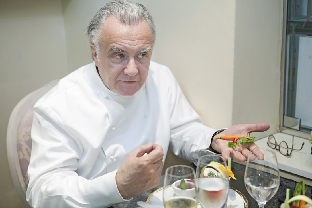 Chef Alain Ducasse in white chef's coat holding a vegetable garnish at his table.