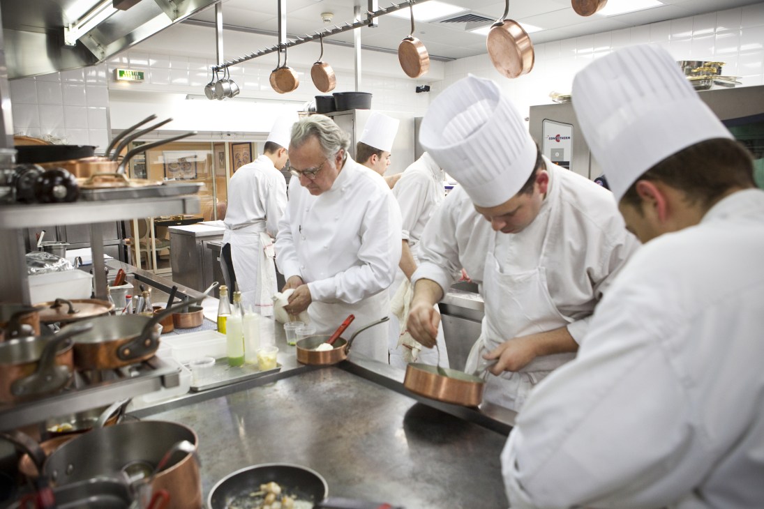 Chef Alain Ducasse works at the stove with kitchen staff in a professional restaurant kitchen.