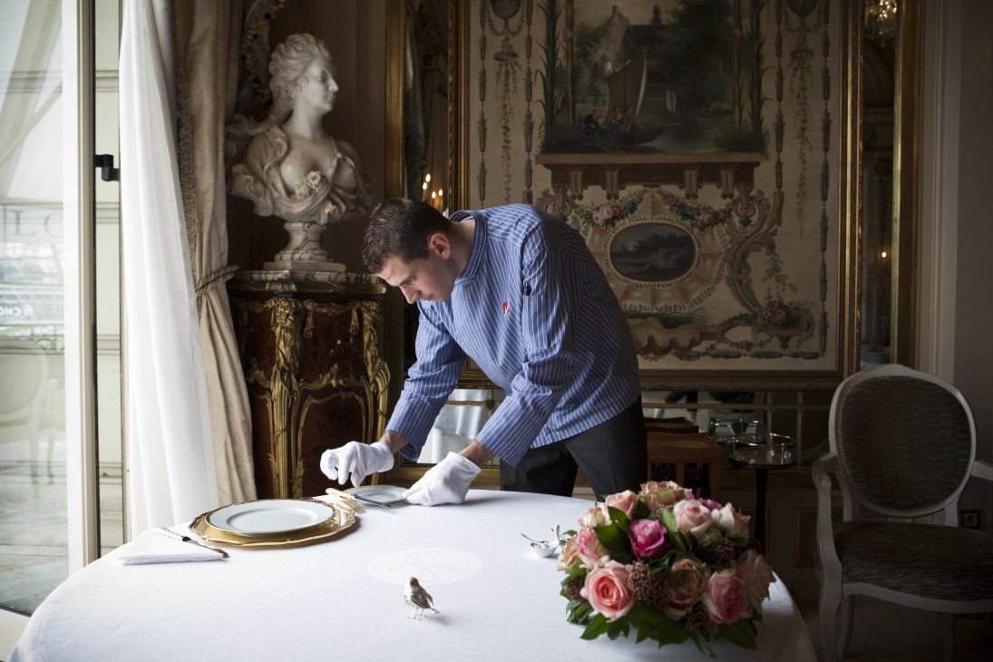 Server in blue pinstriped jacket prepares an ornate dining table at Le Louis XV restaurant.