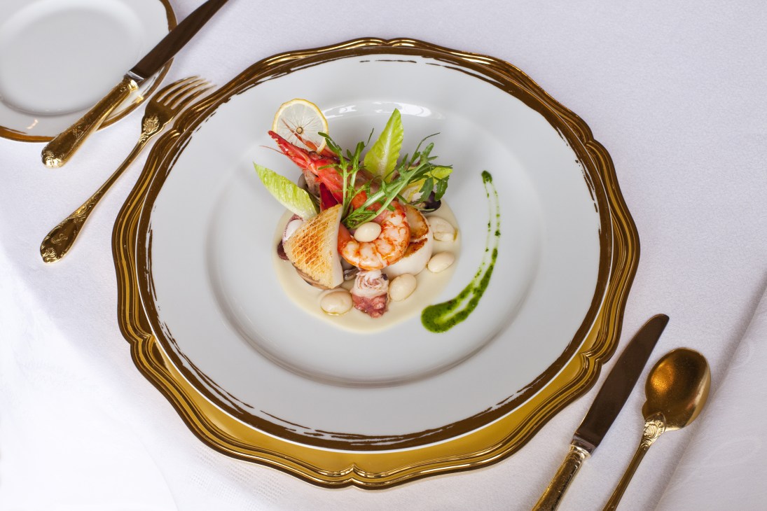 Elegant seafood and coco bean salad plated on fine white china with gold-rimmed rim and ornate gold cutlery.