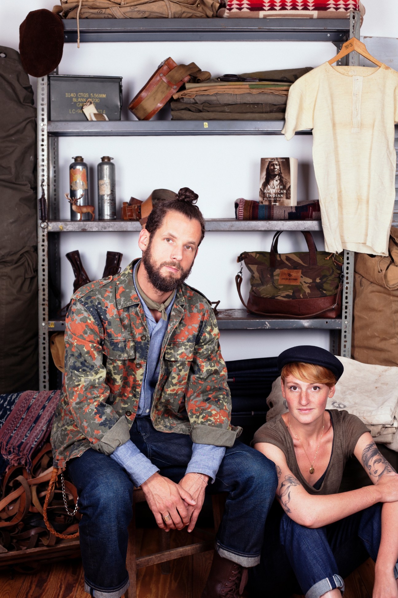 Two people posing in front of industrial shelving displaying military-inspired clothing, bags, and accessories.