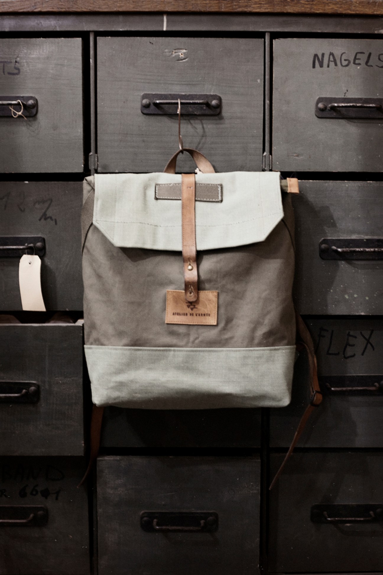 Atelier de l'Armee backpack in cream and grey canvas with brown leather straps hanging on vintage filing cabinet.