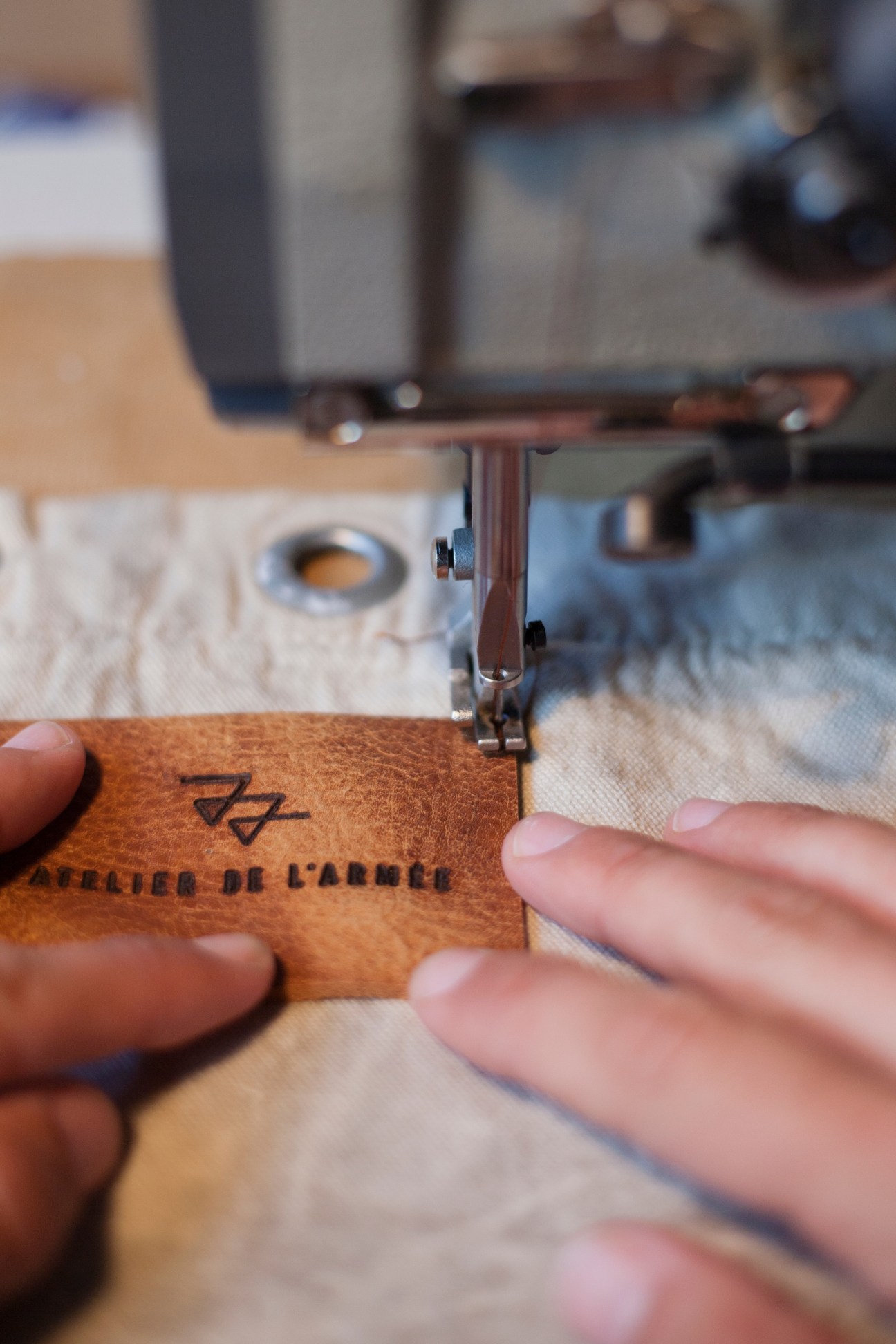 Sewing machine stitching leather label marked 'Atelier de l'Armee' onto fabric at Amsterdam workshop.