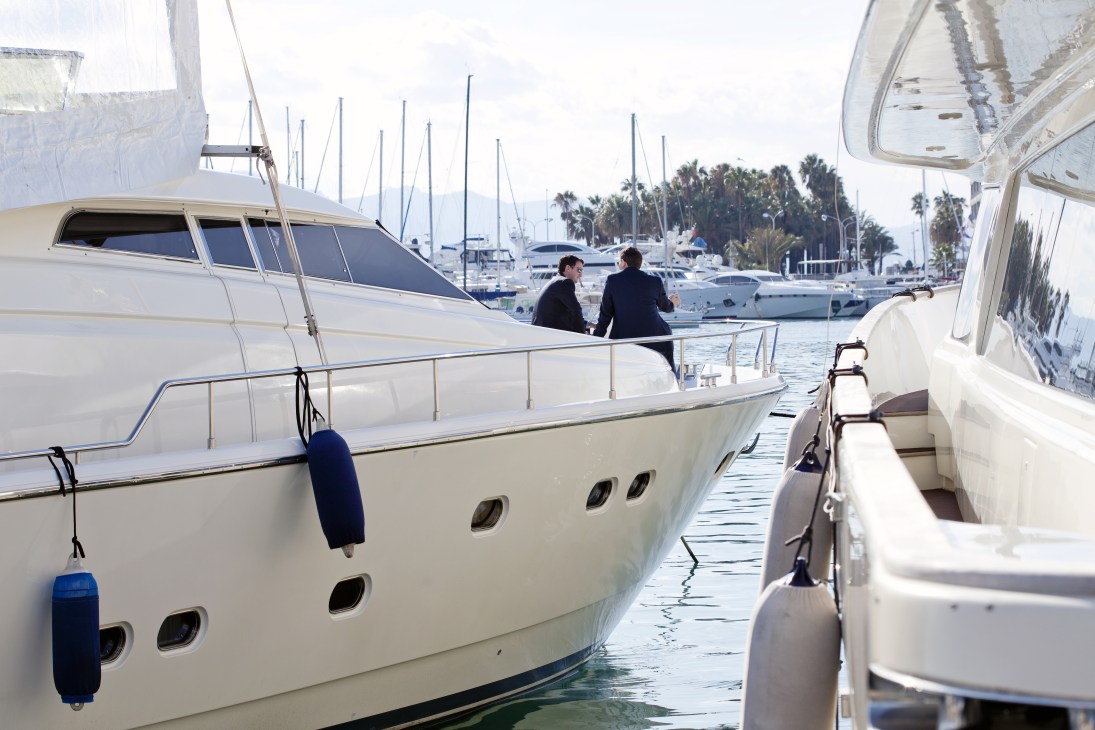 Two people in business attire converse on a luxury yacht moored in a harbor surrounded by sailboats and palm trees.
