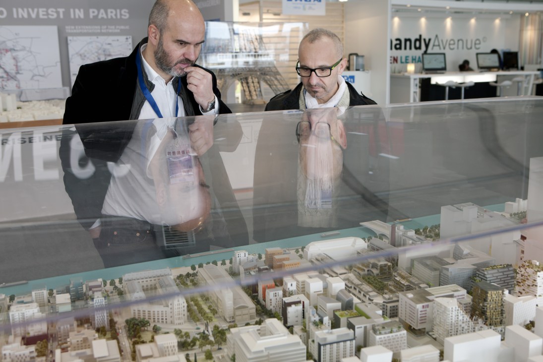 Two men in business attire examine an architectural scale model of a Paris urban development at the MIPIM property fair.
