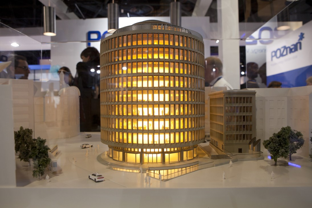 Illuminated architectural model of a cylindrical building at MIPIM real estate fair with exhibition visitors in background.