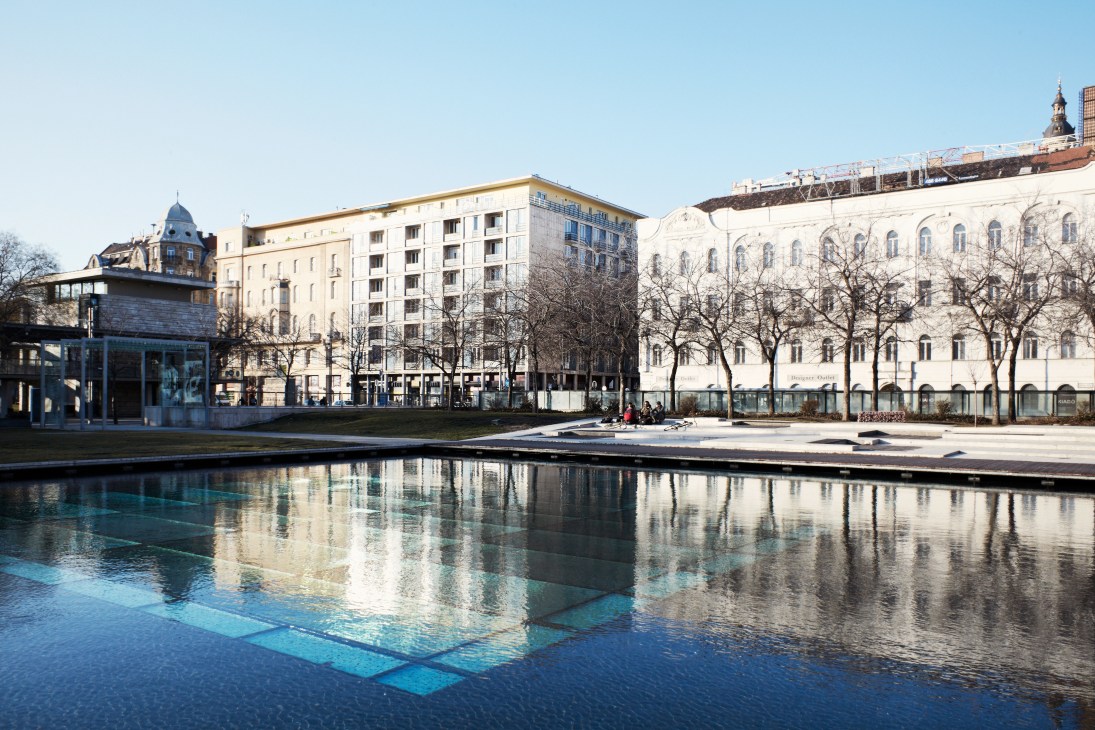 Erzsebet Square in Budapest with modern water feature, historic architecture, and bare trees reflected in still water.