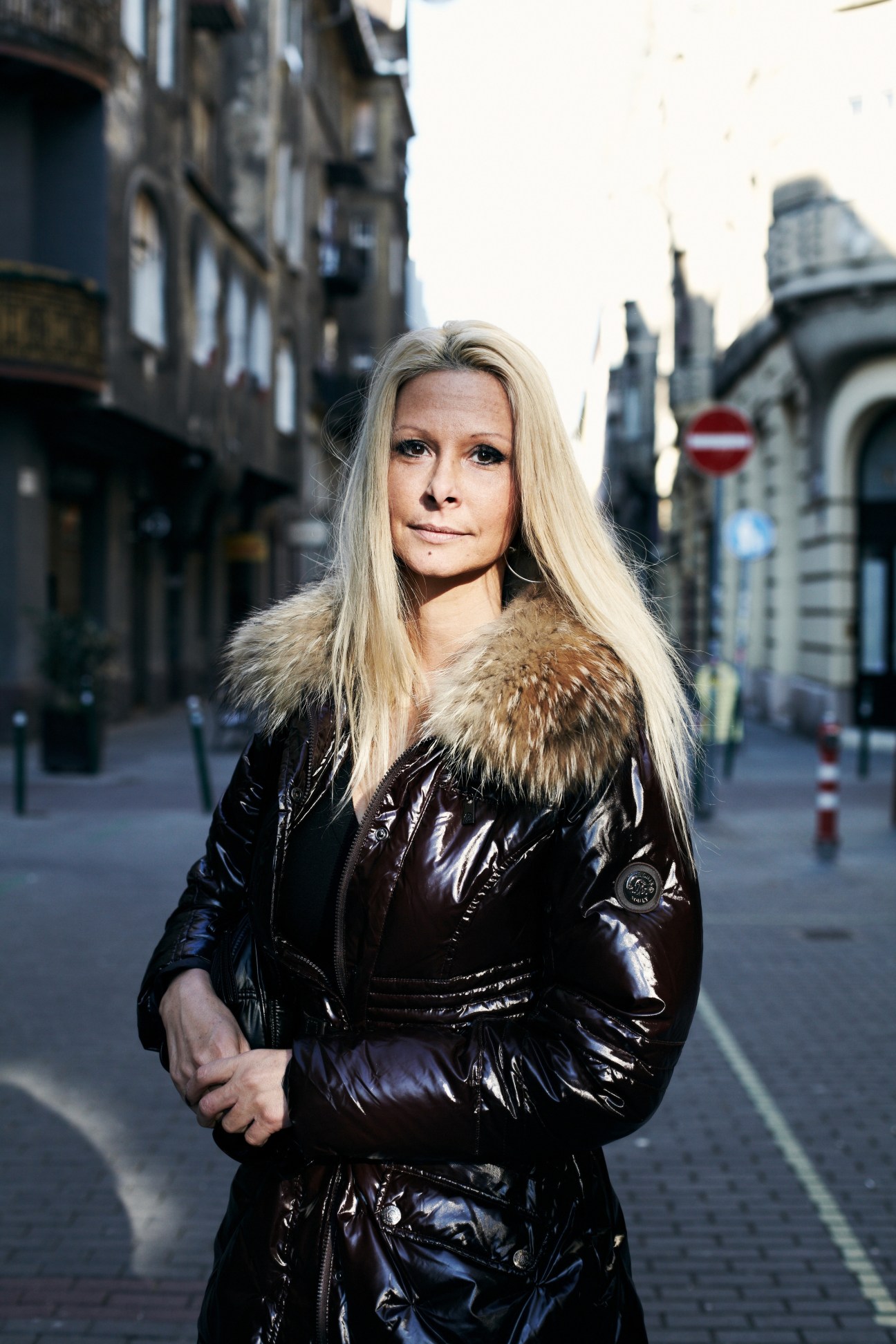 Woman with blonde hair wearing a black puffer jacket standing on a Budapest street with historic buildings in the background.
