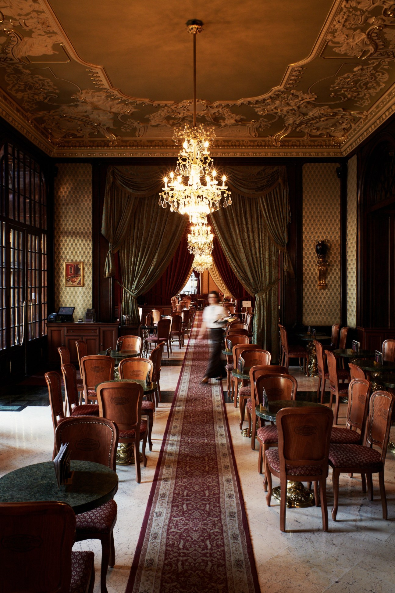 Ornate interior hallway with crystal chandeliers, decorative ceiling, wooden chairs, and draped curtains at Gerbeaud Patisserie.