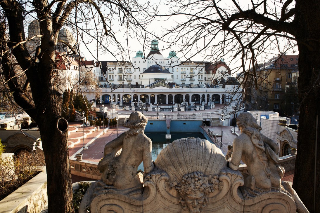 Stone sculptures frame a view of the Gellert Hotel's thermal bath complex and Budapest cityscape with bare trees.