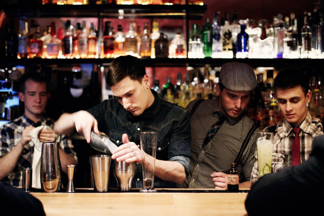 Barman pouring a cocktail at a bar with illuminated bottles on shelves behind him.