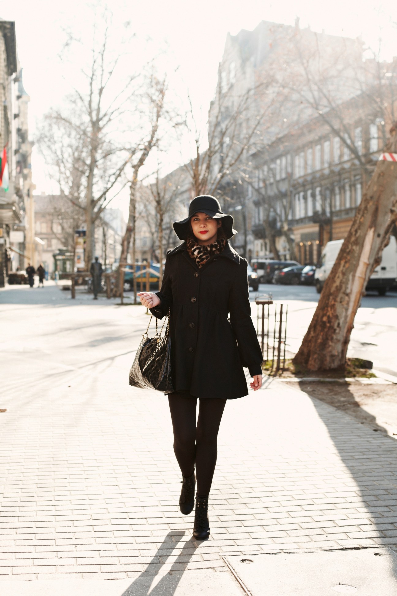 Woman in black coat and hat walking on tree-lined Budapest street carrying a patterned bag.