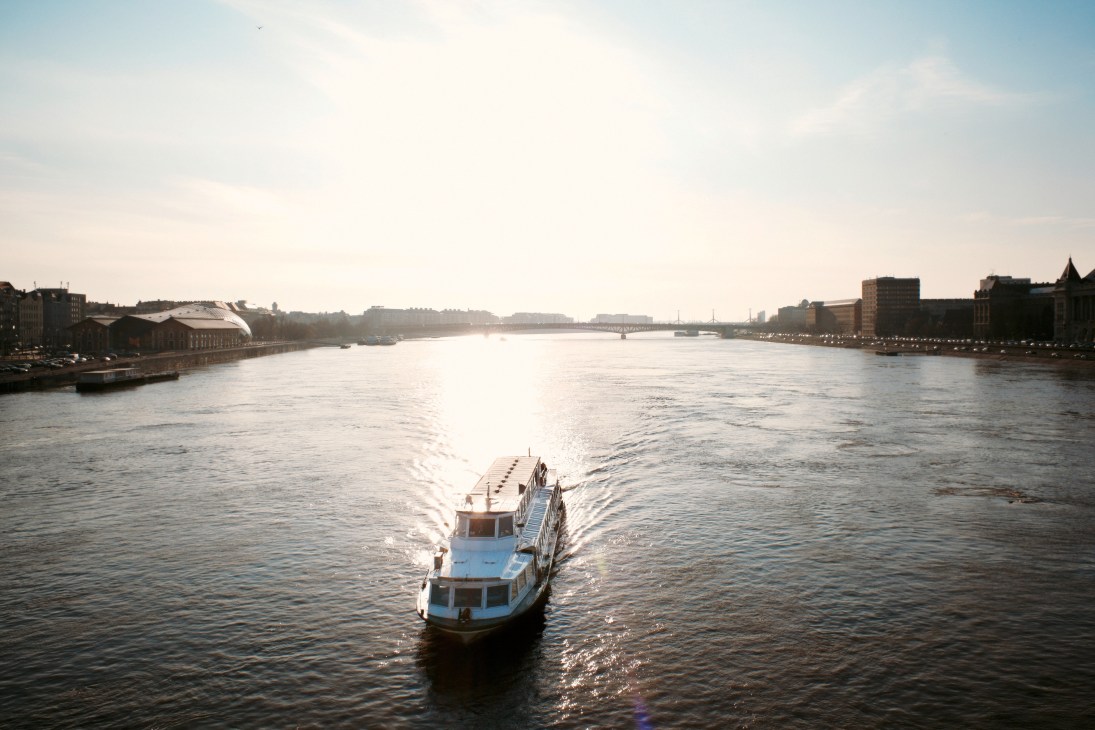A blue and white sightseeing boat travels on the Danube River with Budapest's historic waterfront buildings lining both shores.